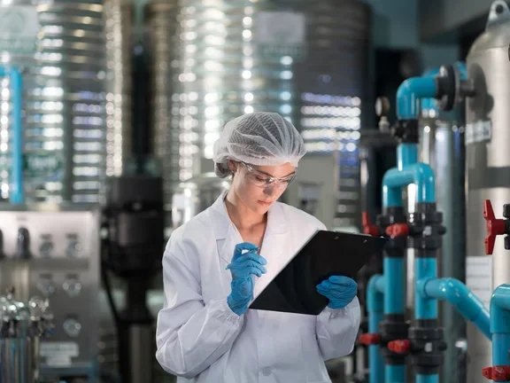 Científica revisando una carpeta en una planta industrial, rodeada de tuberías y tanques.     